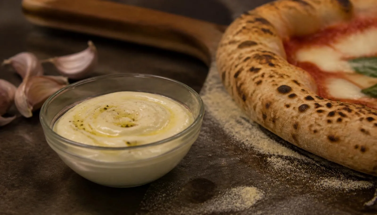 Lille glasskål med hjemmelavet hvidløgsaioli ved siden af den leopardplettede cornicione på en nybagt napolitansk pizza.